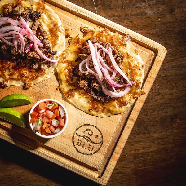 Two Blu tacos with meat, pickled onions, pico de gallo, and lime on a wooden board.