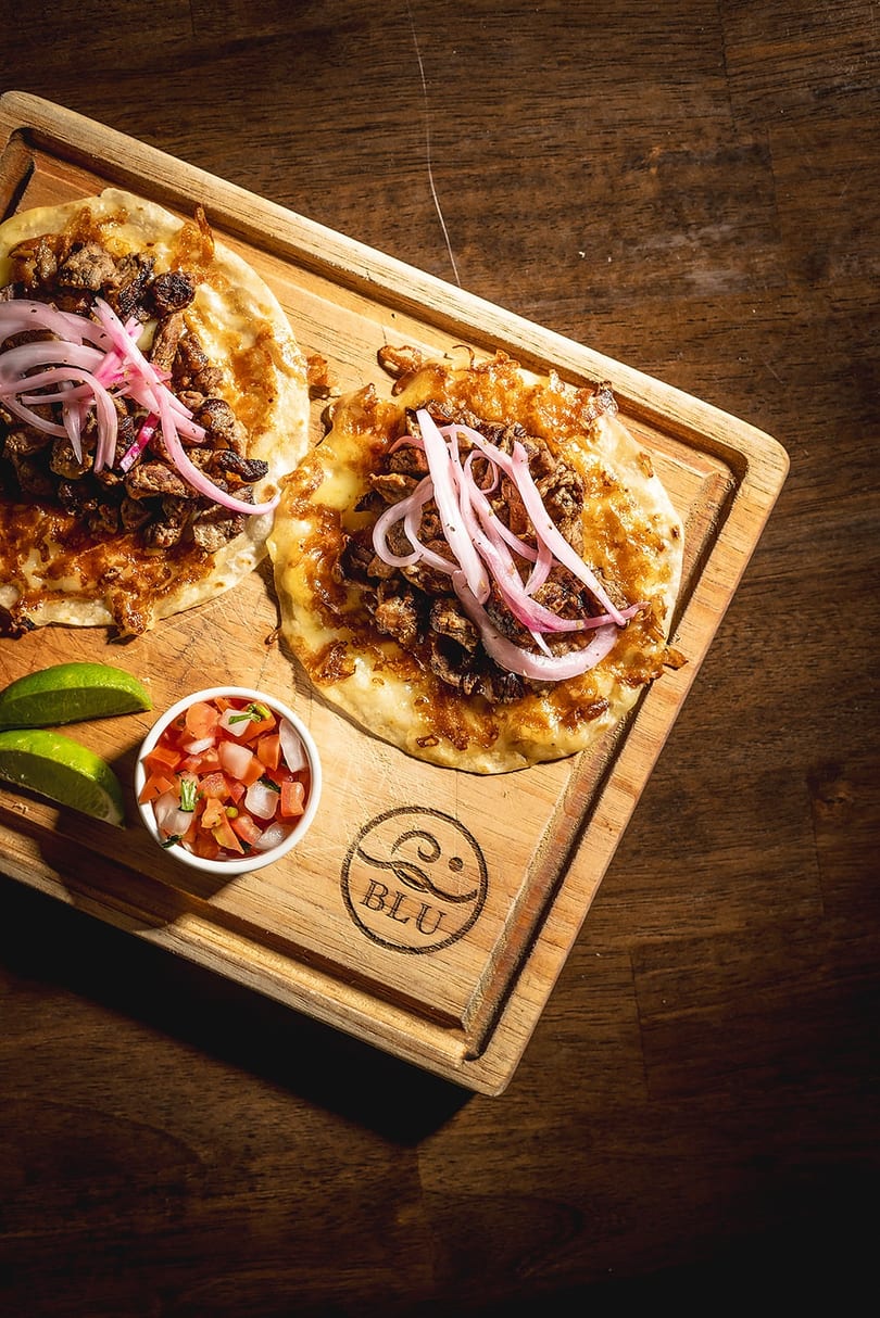 Two Blu tacos with meat, pickled onions, pico de gallo, and lime on a wooden board.