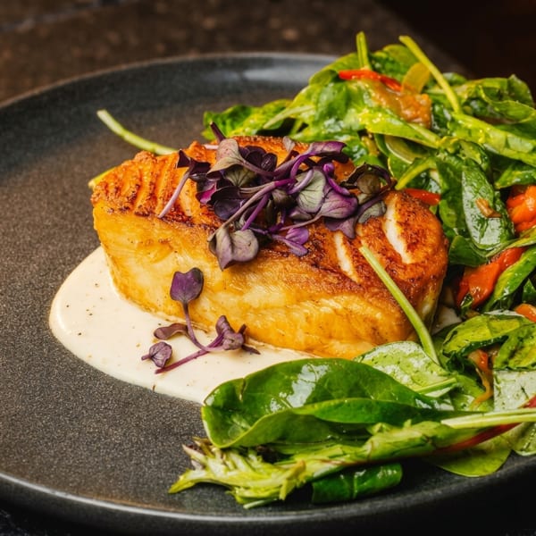 Pan-seared fish with purple microgreens, creamy sauce, and a side of fresh spinach salad.