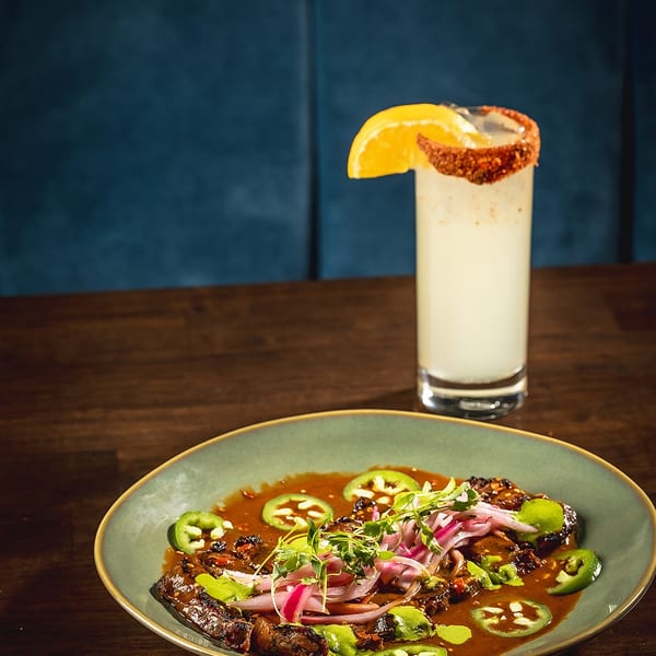 A plate of steak with red onions, jalapeños, and sauce, next to a cocktail with an orange slice.