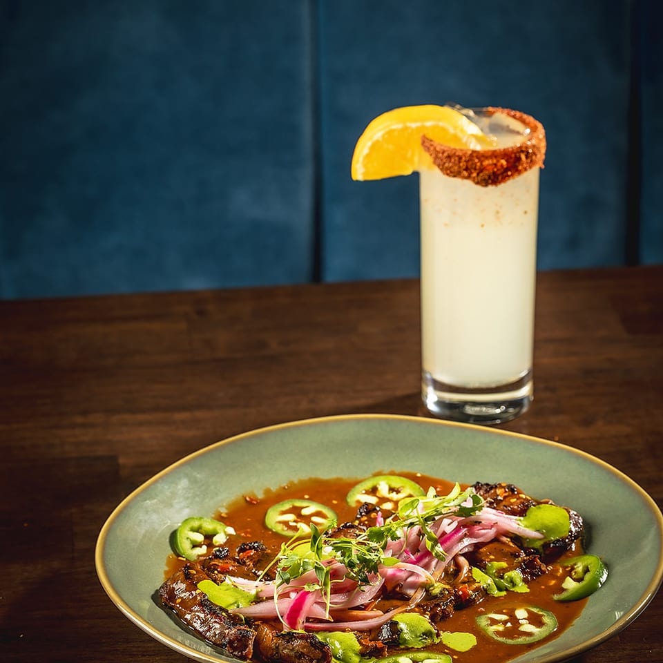 A plate of steak with red onions, jalapeños, and sauce, next to a cocktail with an orange slice.
