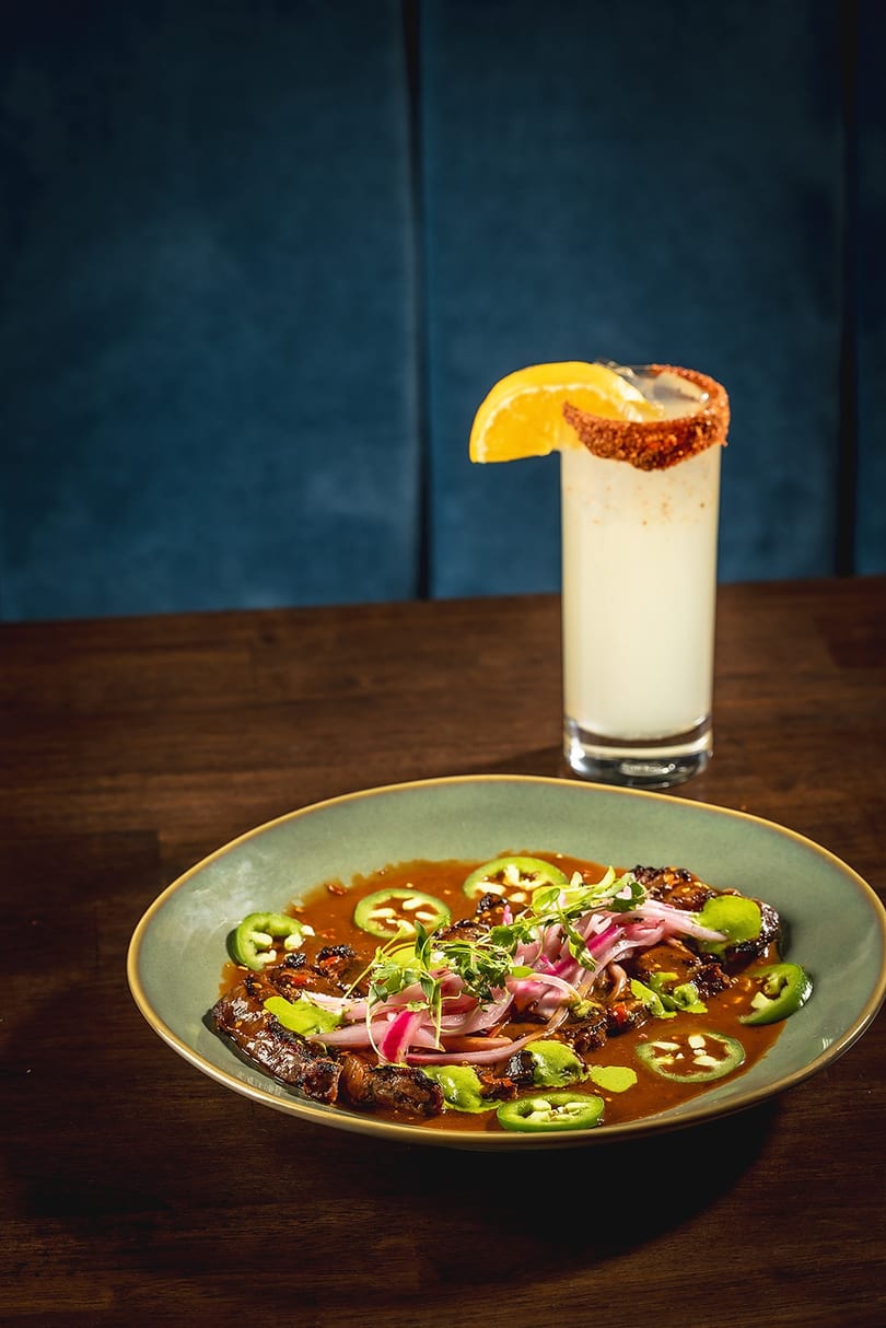 A plate of steak with red onions, jalapeños, and sauce, next to a cocktail with an orange slice.