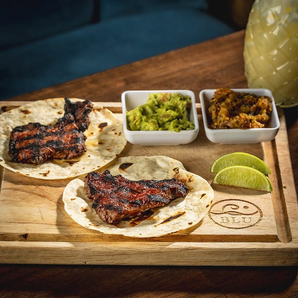 Grilled steak tacos with guacamole, salsa, and lime wedges on a wooden board with a pineapple drink.