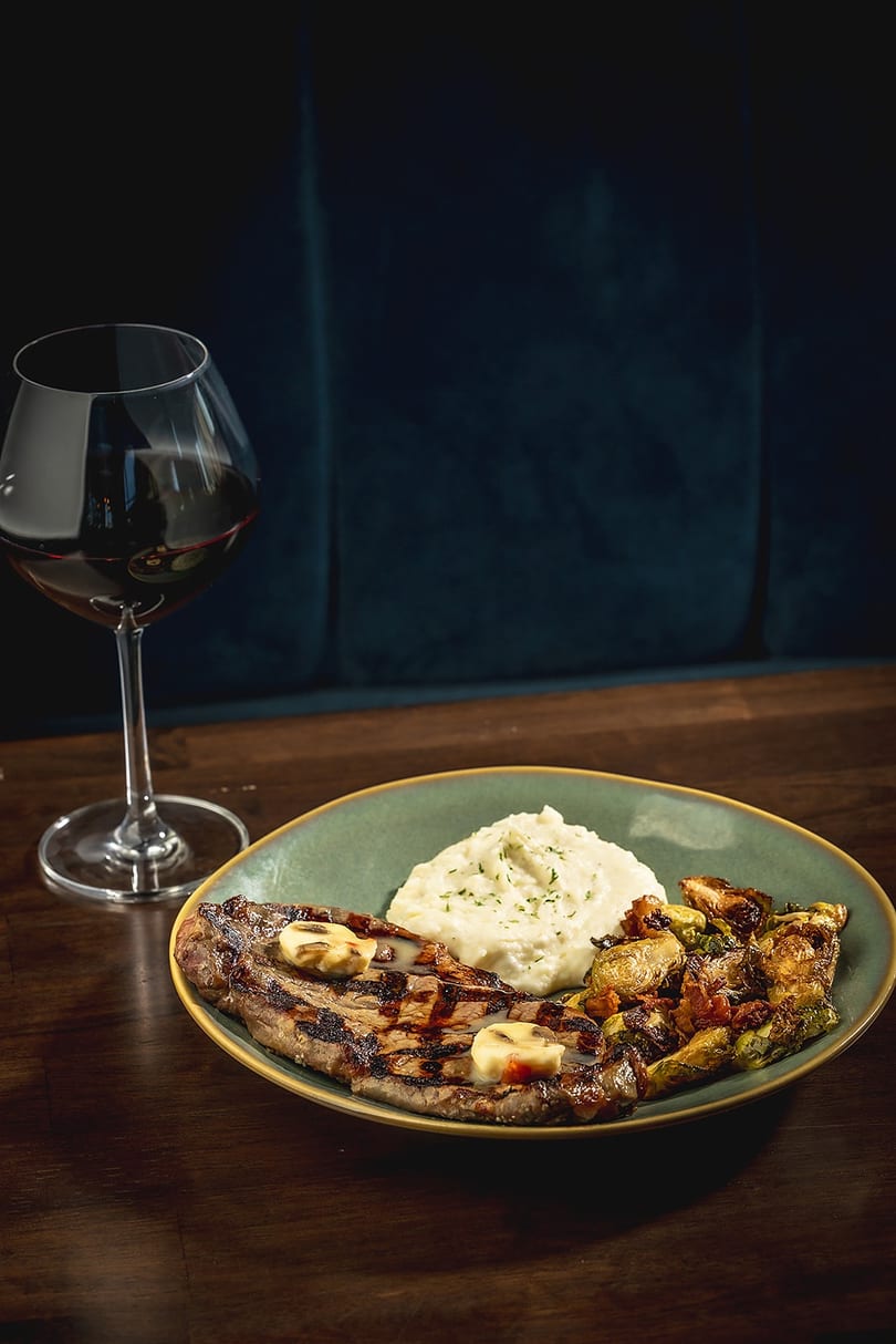 Grilled steak with mashed potatoes and roasted vegetables, next to a glass of red wine.
