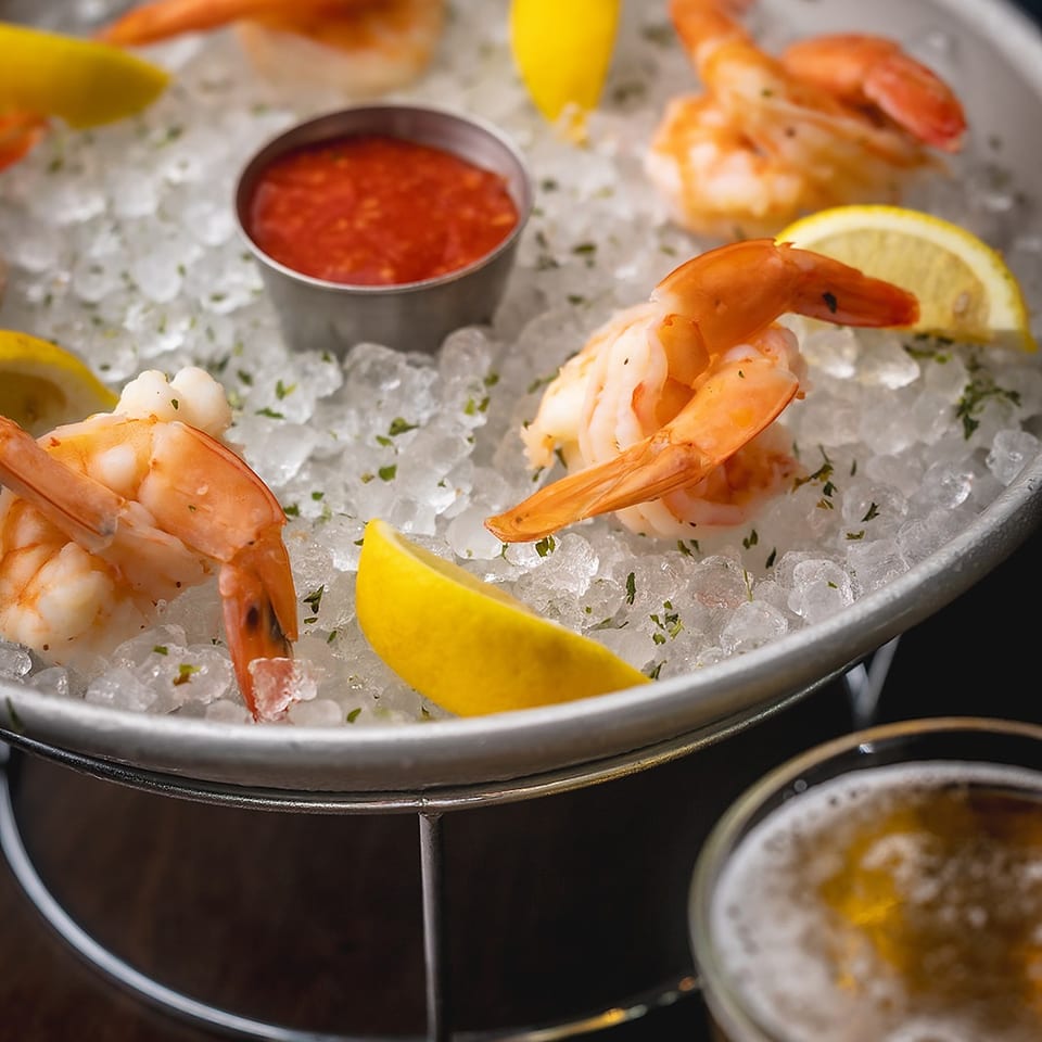 Chilled shrimp cocktail arranged on crushed ice with lemon wedges, dipping sauce, and a beverage on a dark table.