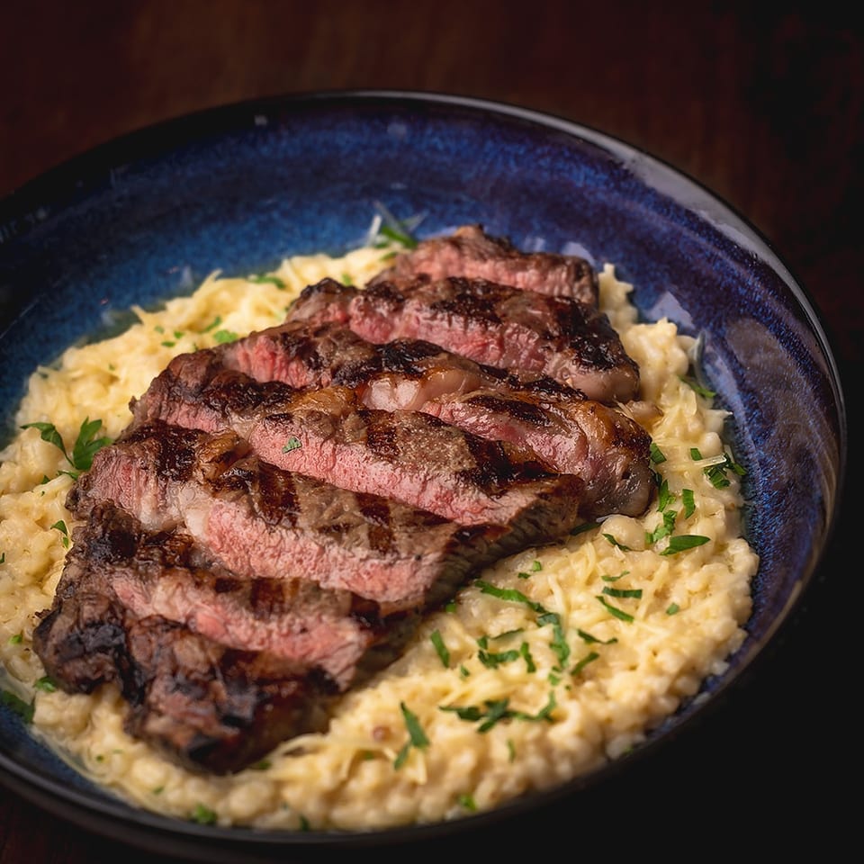 Grilled steak slices arranged over creamy risotto and garnished with fresh parsley in a deep blue bowl.