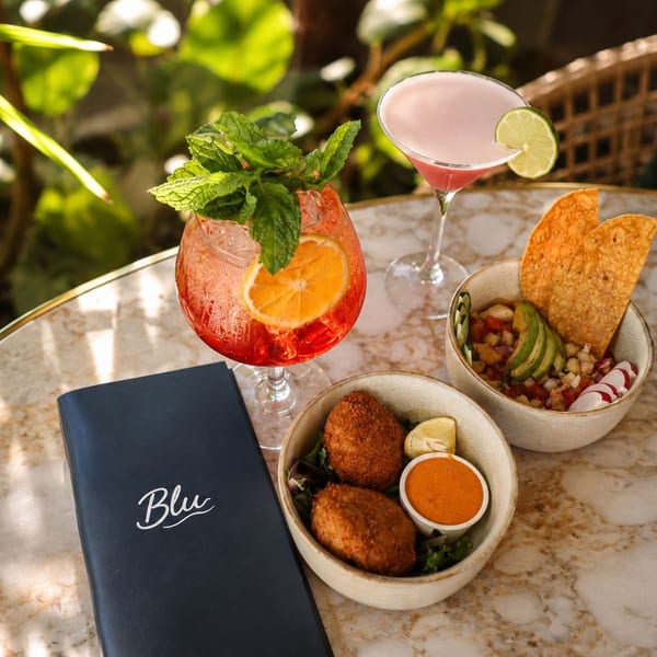 Blu menu, cocktails, and appetizers on a marble table with lush greenery in the background.