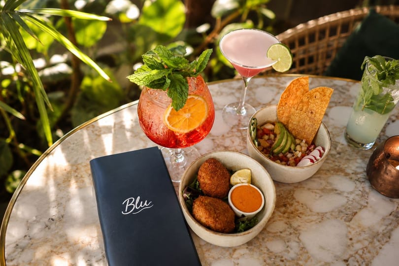 Blu menu, cocktails, and appetizers on a marble table with lush greenery in the background.