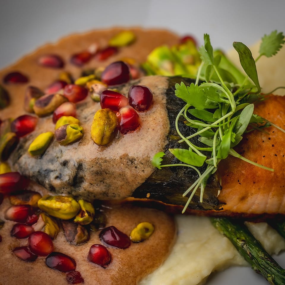 Plated salmon with creamy sauce, pomegranate seeds, pistachios, microgreens, mashed potatoes, and grilled vegetables.
