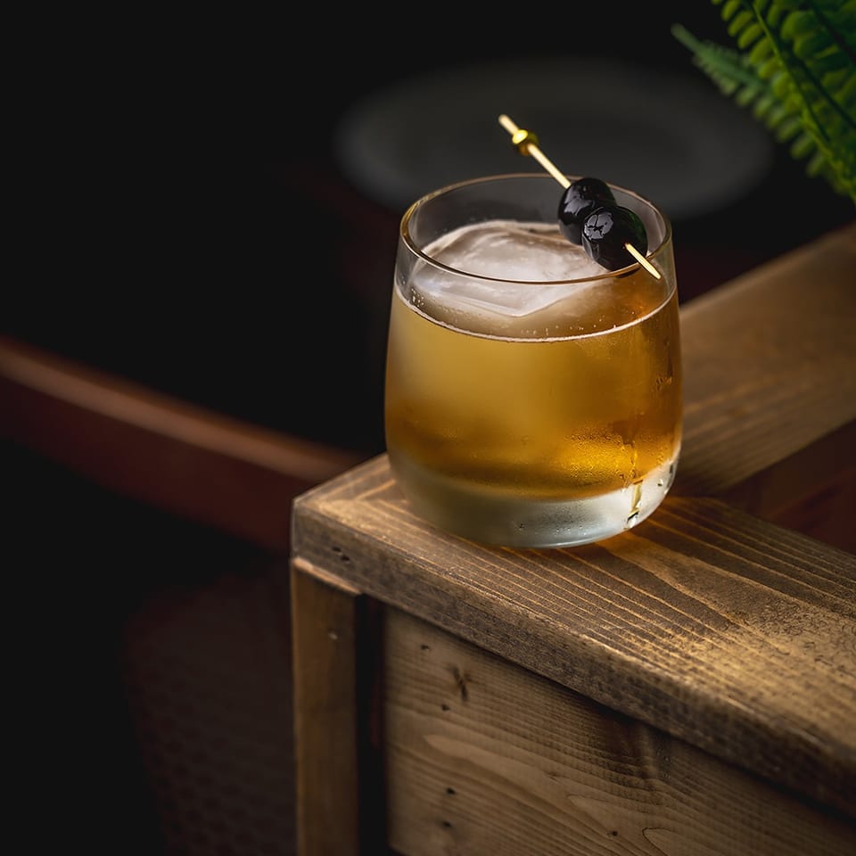 Golden cocktail with foam and two dark cherries on a skewer, resting on a rustic wooden table in a dim setting.