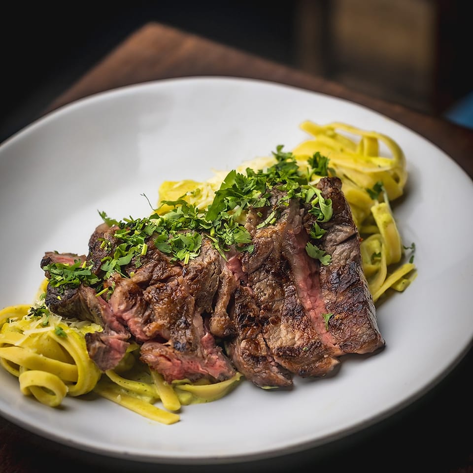 Sliced grilled steak topped with fresh herbs, served over yellow pasta on a white plate.