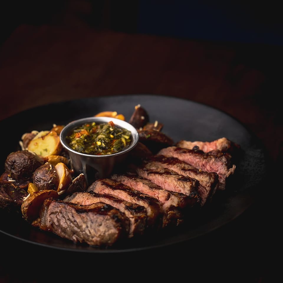 Sliced grilled steak served with roasted potatoes and a bowl of herb sauce on a black plate.
