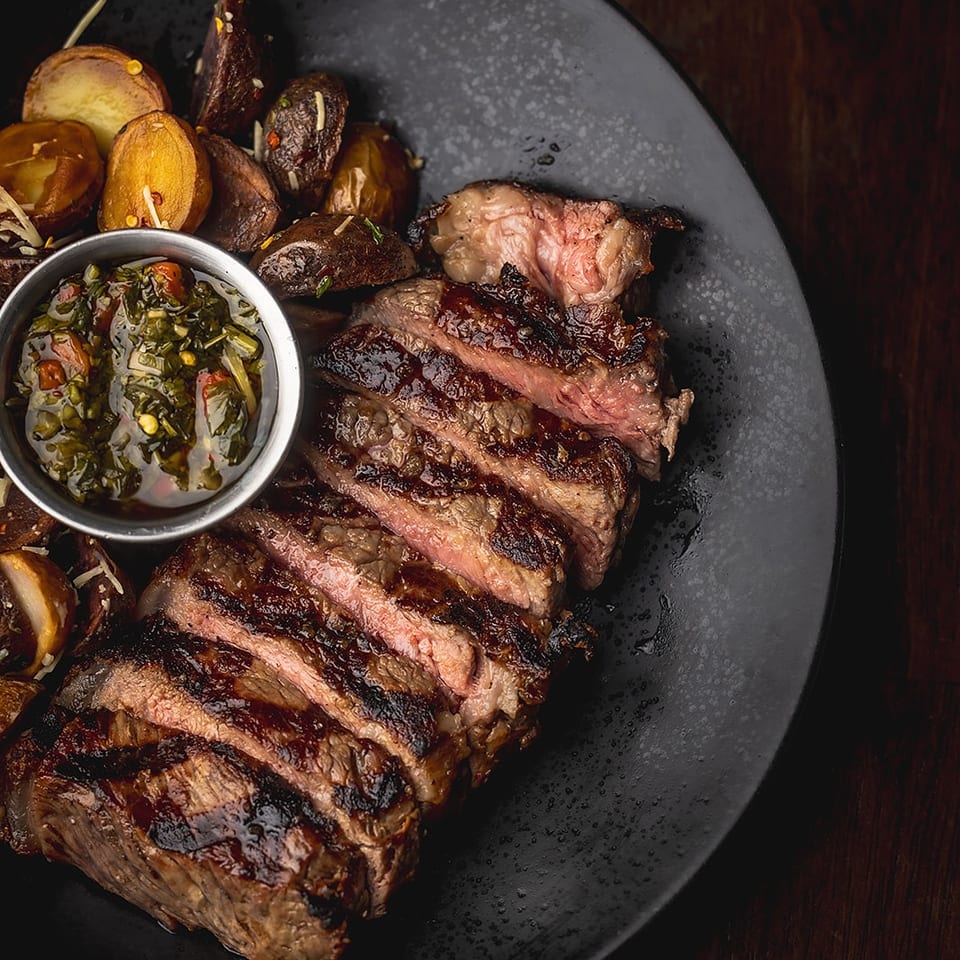 Sliced medium-rare grilled steak with roasted potatoes and a bowl of herb-filled chimichurri sauce on a black plate.