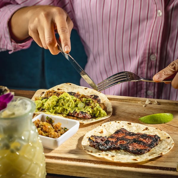 Person in a pink shirt cutting a taco with guacamole and grilled meat on a wooden board.