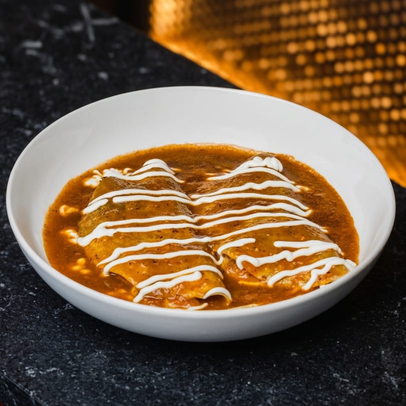 Two enchiladas covered in a rich brown sauce and drizzled with white cream, served in a white bowl.