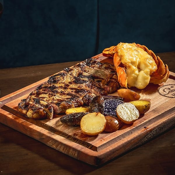 Grilled steak and lobster tail with roasted potatoes on a wooden board, with a glass of red wine.