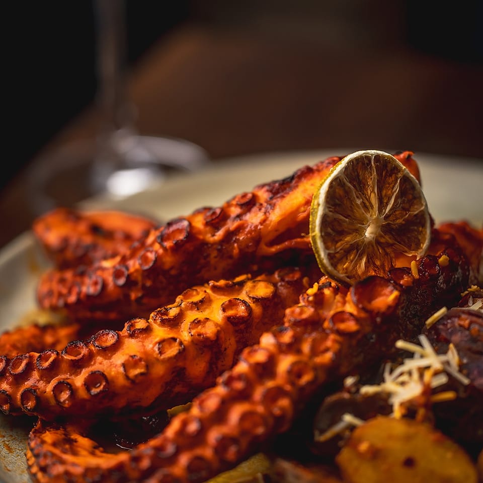 Grilled octopus tentacles with a dried lemon slice and roasted vegetables on a plate, paired with a wine glass.