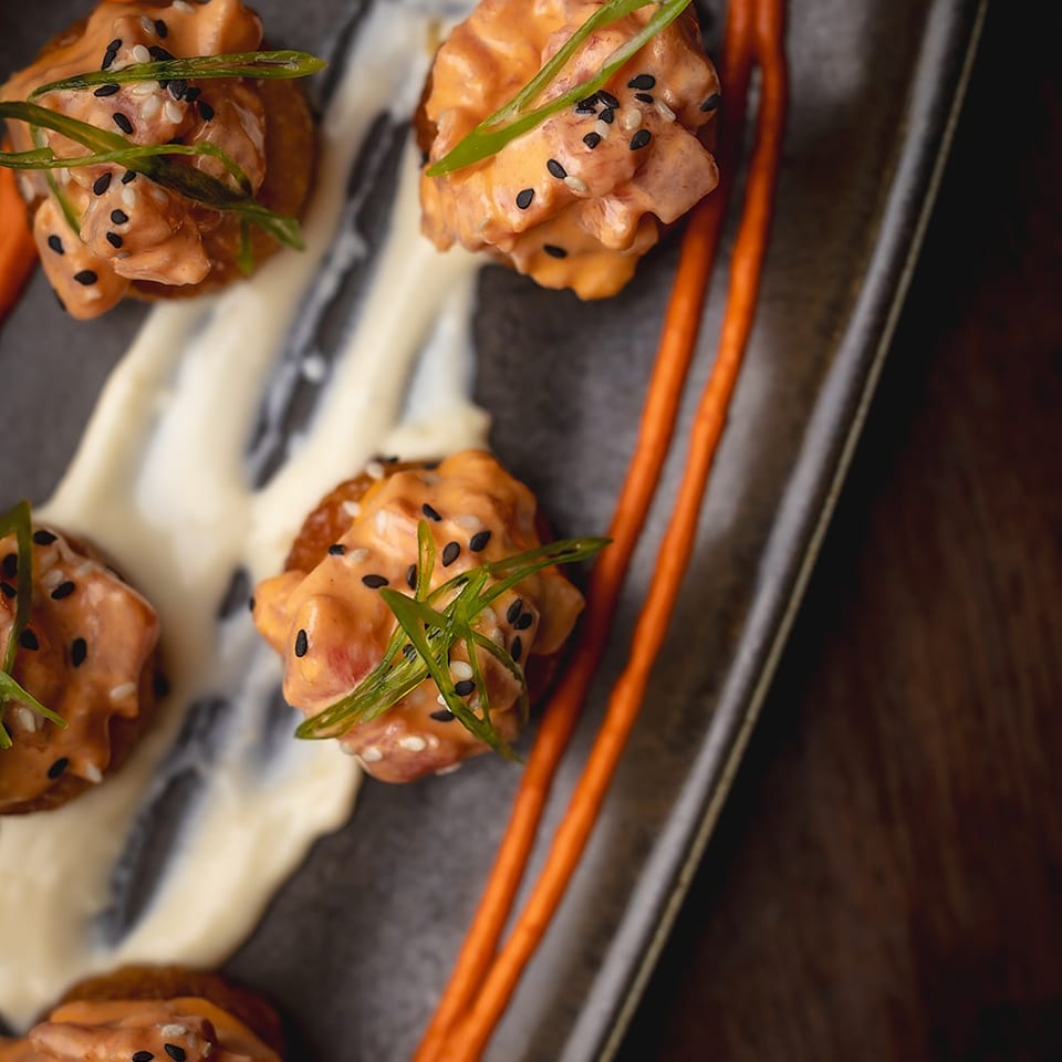Plated orange-glazed bite-sized appetizers topped with black and white sesame seeds and scallions, drizzled with cream sauce and orange sauce.