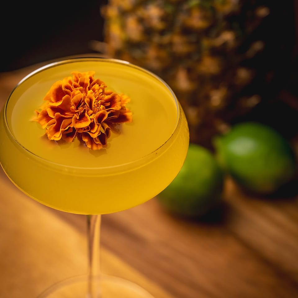 Golden cocktail garnished with an orange marigold flower, with a blurred pineapple and lime in the warmly lit background.