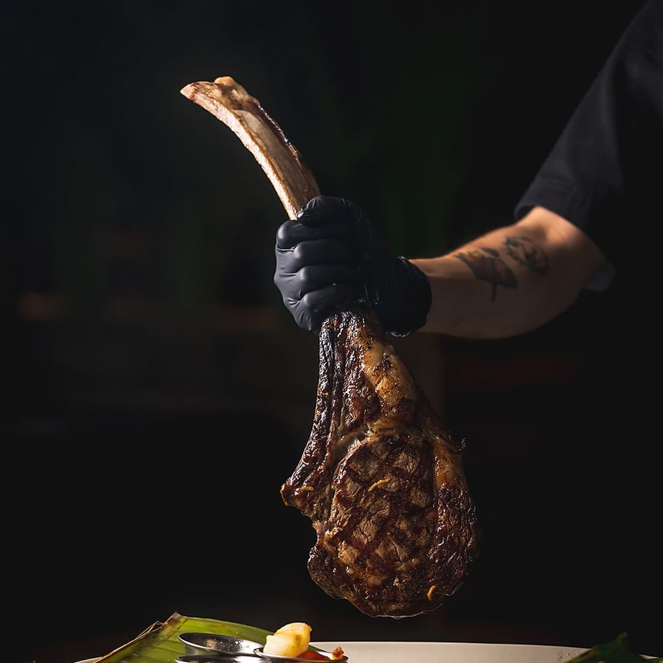 Gloved server holds a grilled tomahawk steak above a banana-leaf-lined plate with colorful vegetables.