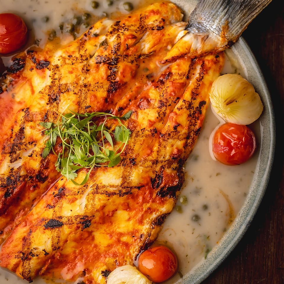 Grilled fish fillet with crispy skin, creamy herb sauce, roasted tomatoes and onions, and fresh greens on a ceramic plate.