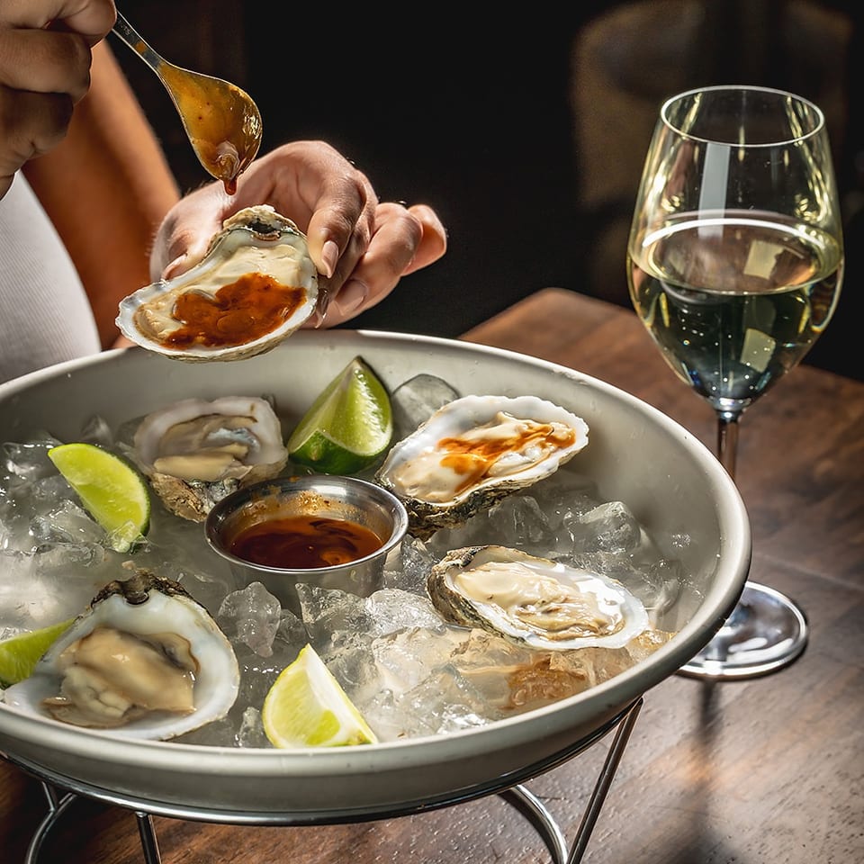Person drizzles sauce over oysters on ice with lime wedges, dipping sauce, and a glass of white wine.