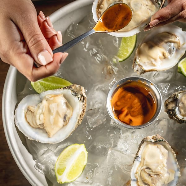 Hands spooning sauce onto a fresh oyster on a bed of ice with lime wedges.
