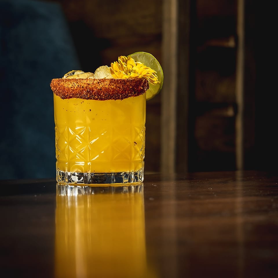 Golden cocktail in a patterned glass with a chili-salt rim, lime wedge, edible flower, and ice on a dimly lit bar.