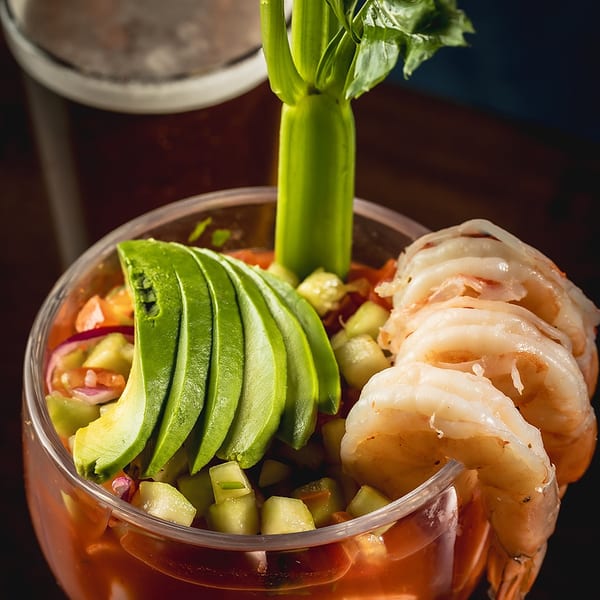 Shrimp cocktail with avocado, cucumber, and celery in a glass, with a beer in the background.