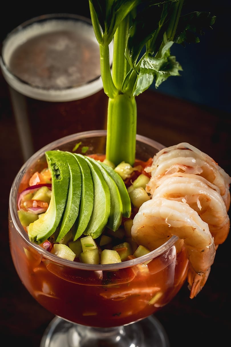 Shrimp cocktail with avocado, cucumber, and celery in a glass, with a beer in the background.