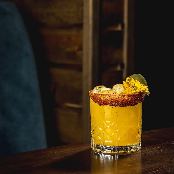 Yellow cocktail with a spiced rim and a yellow flower garnish on a dark wooden bar.