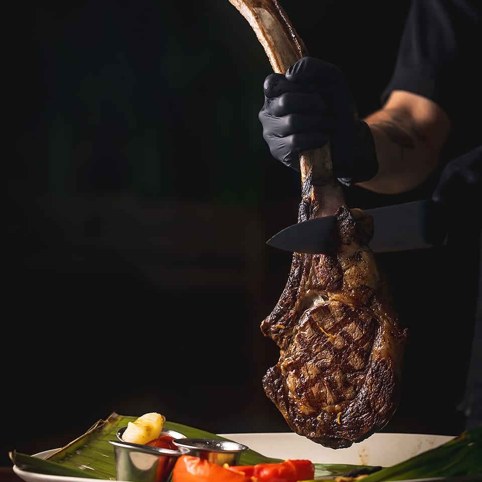 Gloved server slices roasted meat from a large bone over a banana-leaf-lined plate with vegetables and dipping sauces.