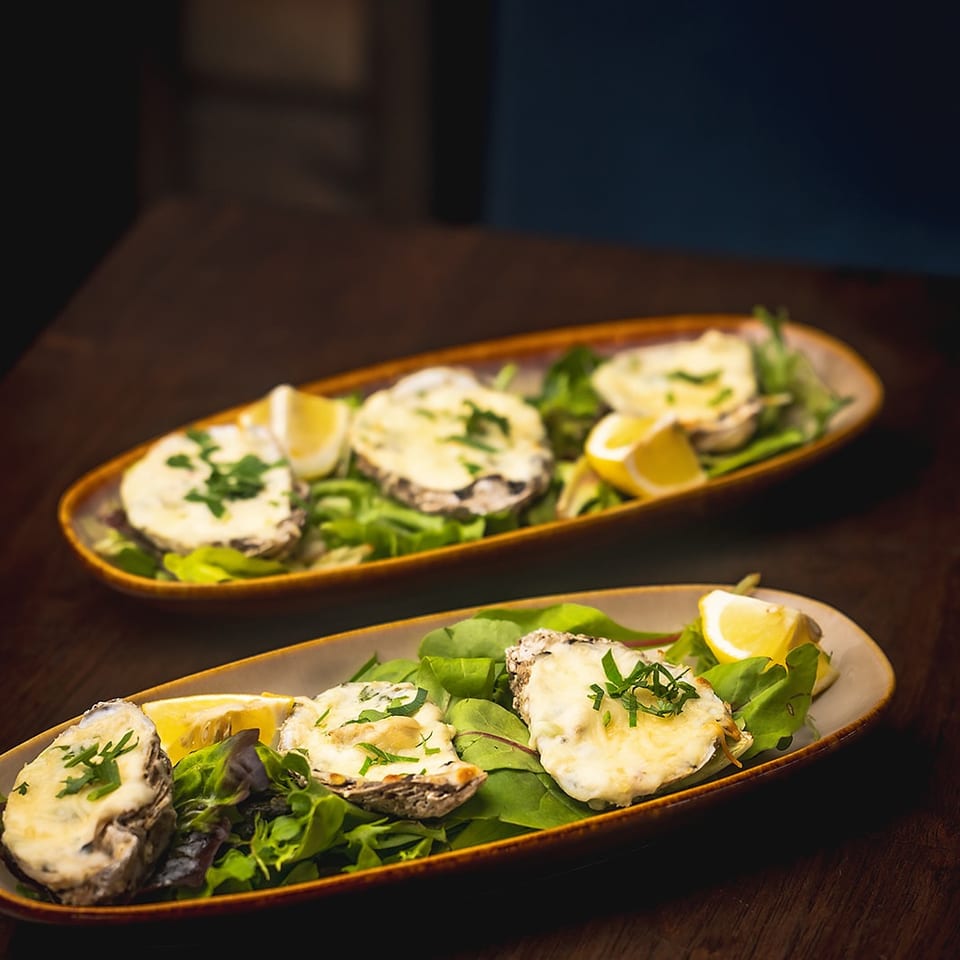 Two long platters hold baked oysters topped with herbs, served over leafy greens with lemon wedges on a wooden table.
