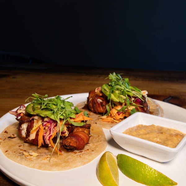 Two bacon-wrapped tacos with slaw, avocado, and microgreens, served with lime wedges and sauce.