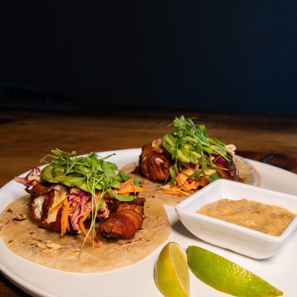 Two bacon-wrapped tacos with slaw, avocado, and microgreens, served with lime wedges and sauce.