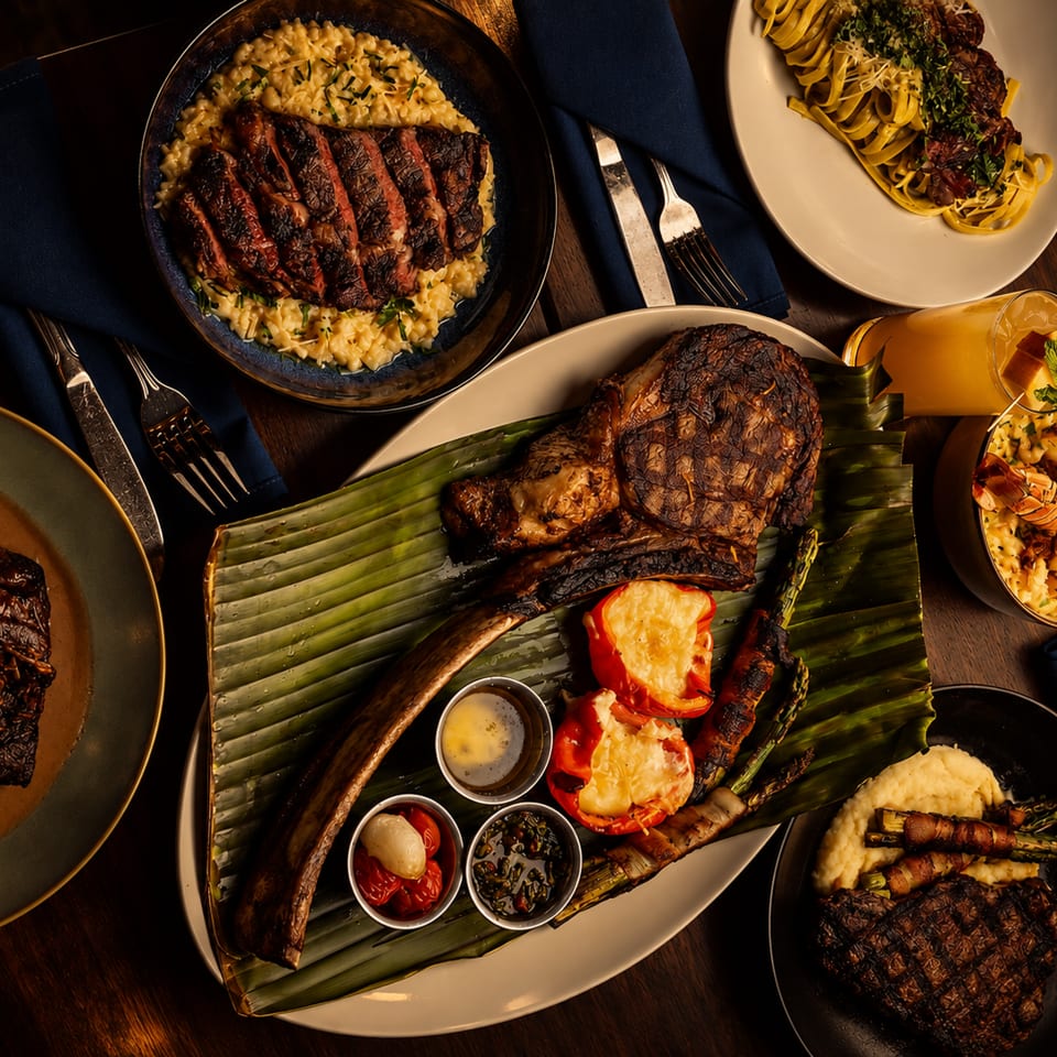 Overhead view of a lavish steakhouse spread with grilled meats, lobster, pasta, mashed potatoes, vegetables, and cocktails on a wooden table.