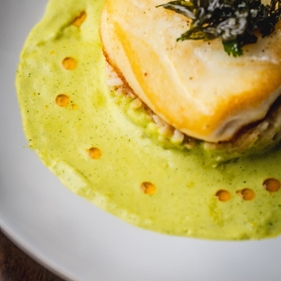 Seared fish topped with dark leafy greens, served over creamy green herb sauce on a white plate.
