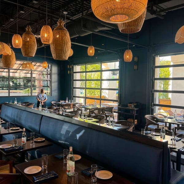 Restaurant interior with blue booth seating, dark tables, wicker light fixtures, and large windows.