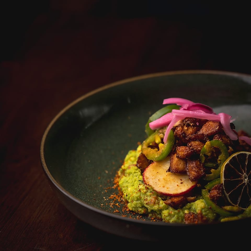 A dark plate holds seasoned roasted meat with guacamole, pickled onions, jalapeños, radishes, and charred lime.