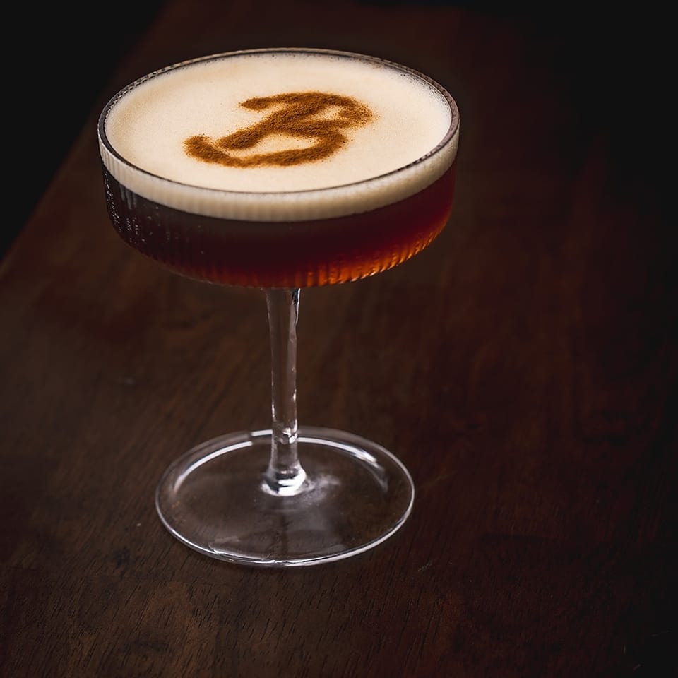 Dark cocktail with a creamy foam top and cinnamon garnish served in a coupe glass on a wooden bar.