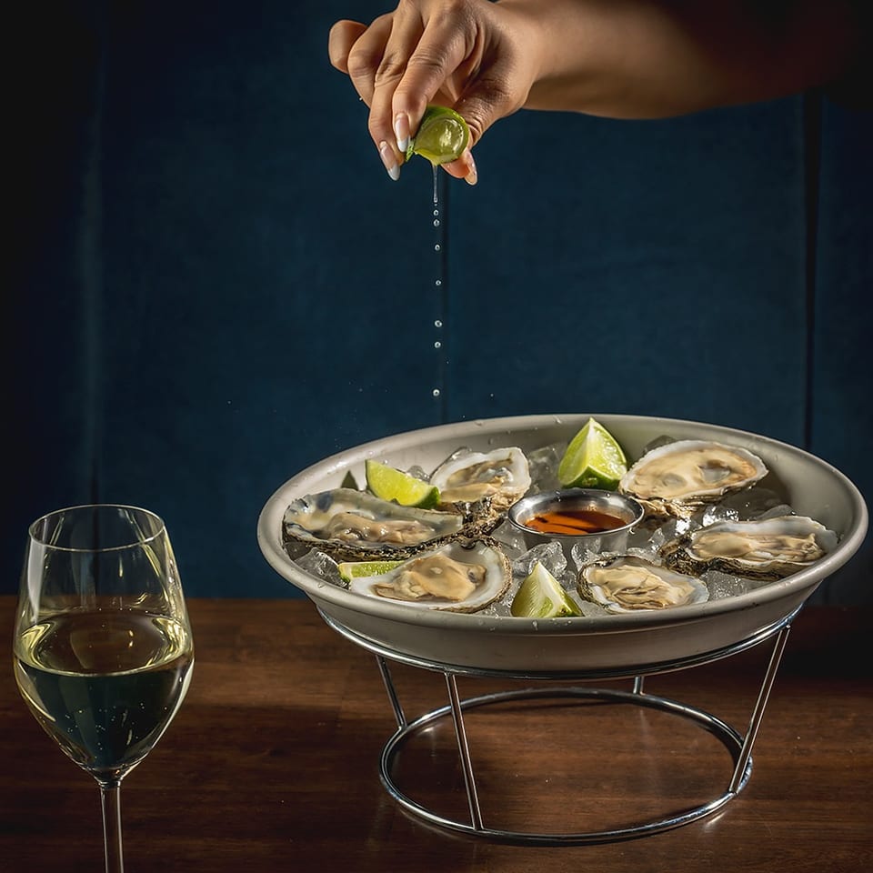 Hand squeezing lime over oysters on ice with a glass of white wine on a wooden table.