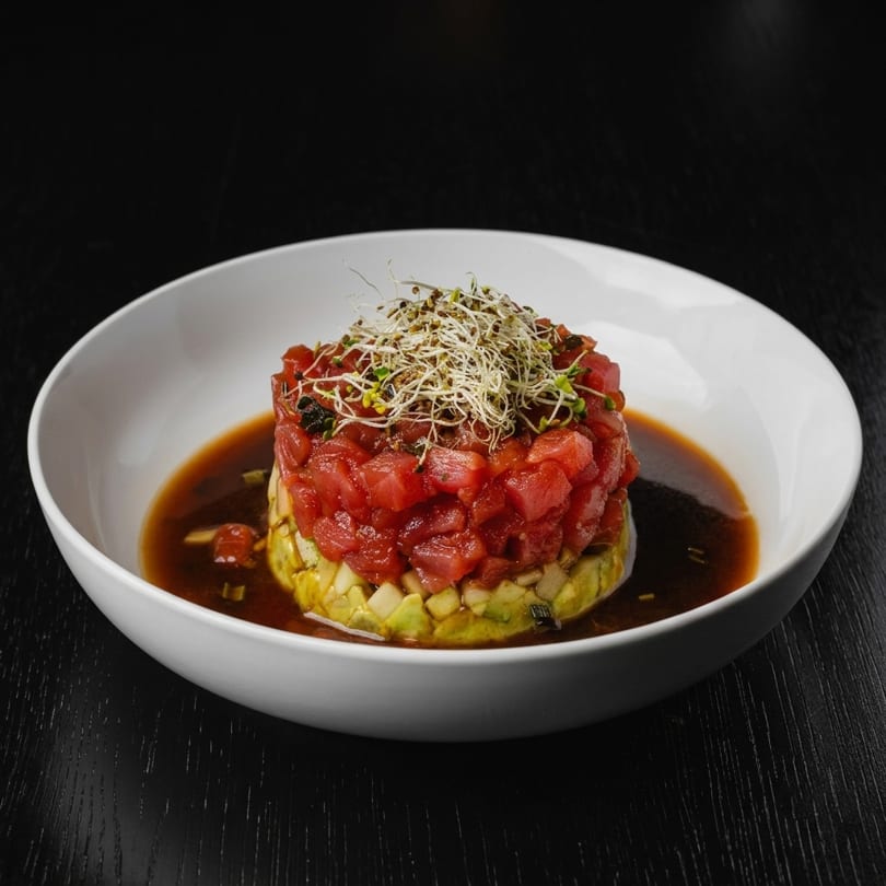 Tuna and avocado tartare with sprouts in a white bowl with dark sauce.