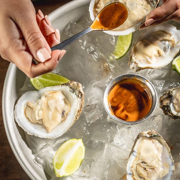 Hands spooning sauce onto fresh oysters on a bed of ice with lime wedges and dipping sauce.