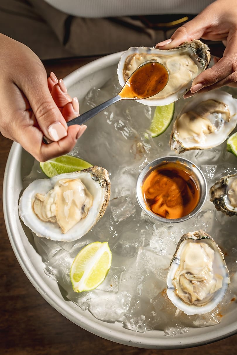 Hands spooning sauce onto fresh oysters on a bed of ice with lime wedges and dipping sauce.