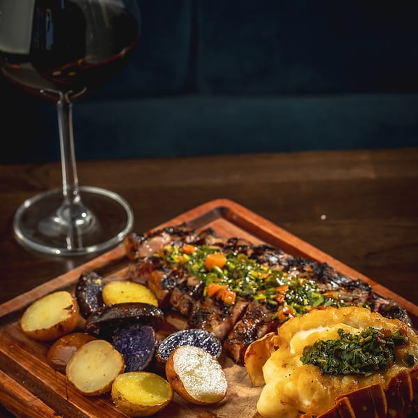 Grilled steak with chimichurri, roasted potatoes, and lobster tail on a wooden board with red wine.