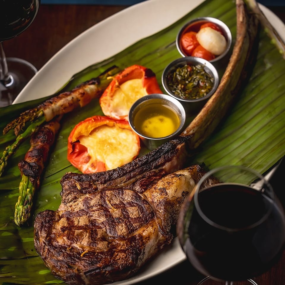 Grilled steak with asparagus, roasted tomatoes, dipping sauces, and wine served on a banana leaf-lined plate.