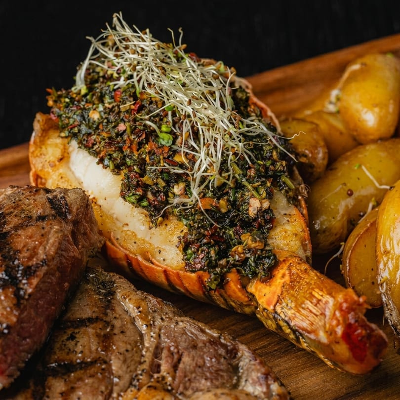 Grilled lobster tail with green herb topping and sprouts, served with steak and potatoes.